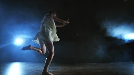 A-young-female-ballerina-barefoot-jumps-on-stage-and-moves-in-slow-motion-in-a-loose-white-dress.